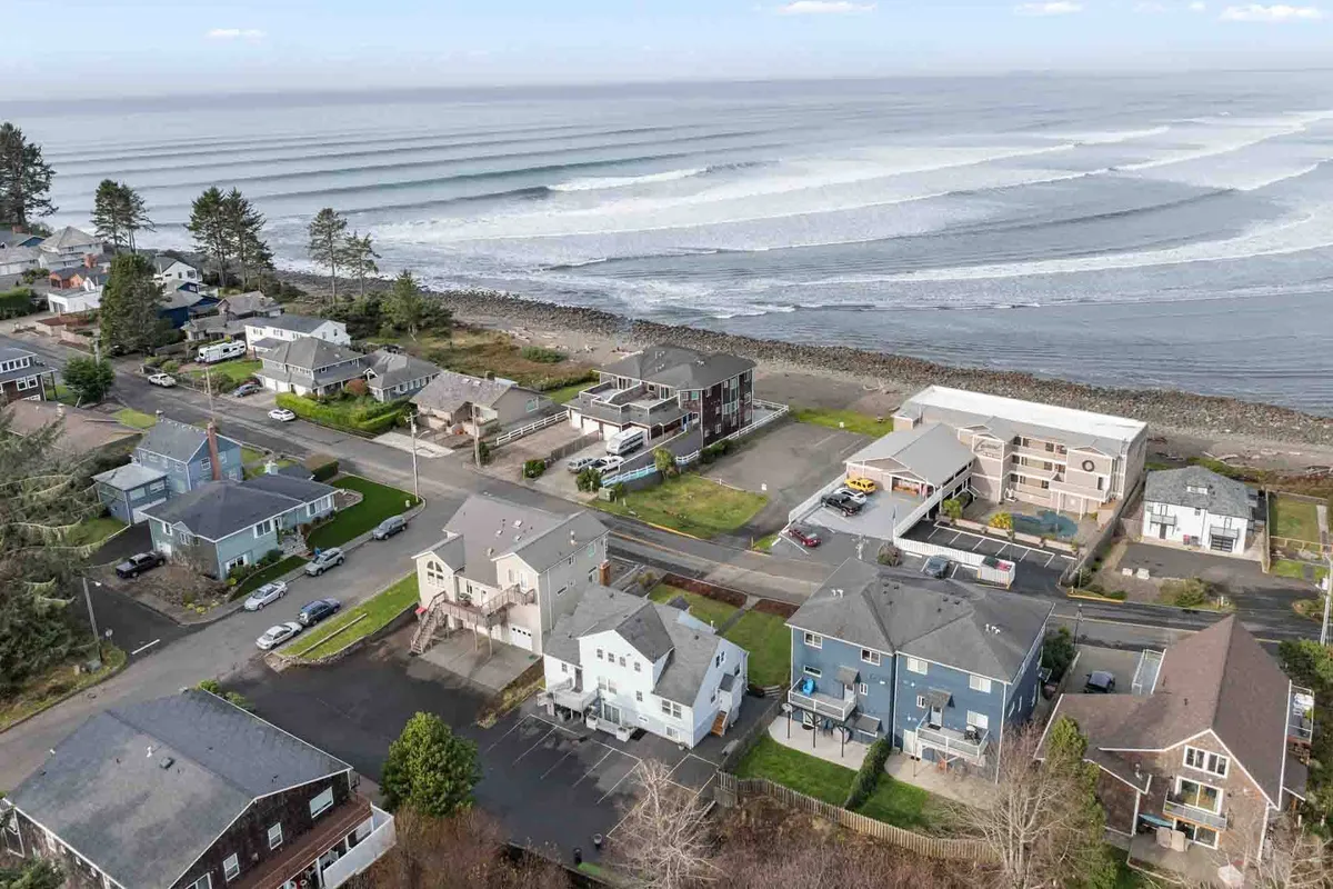 A sweeping view of the North Coast beachfront, with gentle ocean waves rolling onto the sand, a row of beachfront properties in the distance, coastal trees lining the horizon, and winding roadways visible. The scene is tranquil, with early or late sunlight casting a soft glow, evoking the beauty of seaside exploration and outdoor recreation.