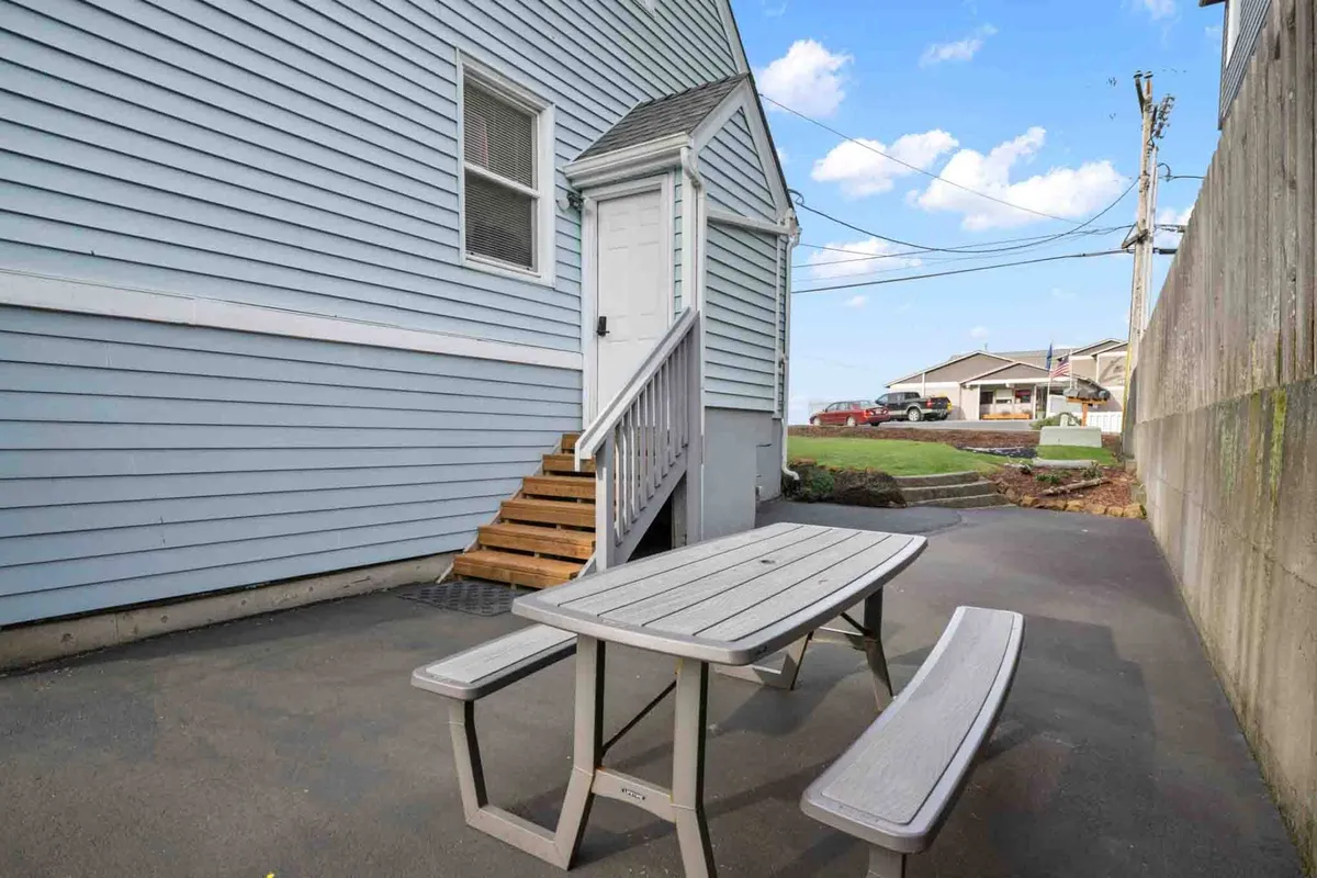 An outdoor dining area featuring a table and benches next to the house, with stairs leading to the entrance and a cheery blue house exterior. The scene is set for outdoor meals or socializing, with space for groups to gather under the sun or stars.