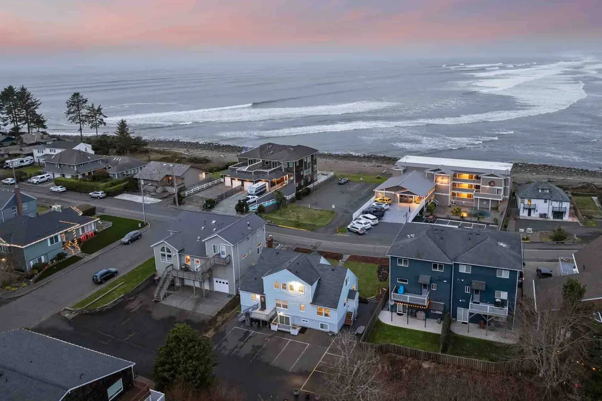 A scenic view of ocean waves rolling towards a sandy shore with houses lining the coast and a vibrant sunset sky overhead. The image captures the essence of family-friendly beach activities such as sandcastle building, surfing, and taking walks along the water.