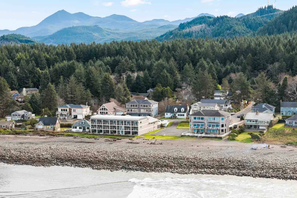 A sweeping view of beachfront properties nestled against a mountain backdrop, with a rocky shoreline in front. The scene is vibrant with activity: families gathering, people beachcombing, and hikers exploring the nearby mountains. The light suggests either sunrise or sunset, casting a relaxed and inviting glow over the landscape.