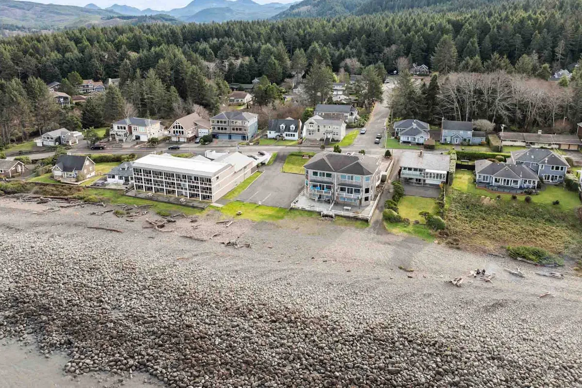 This inviting exterior view features beachfront houses bordered by a rocky shoreline, with a lush forest backdrop and distant mountains. People are engaged in beachcombing and walking along the shore, evoking a sense of connection with nature and opportunities for outdoor exploration.