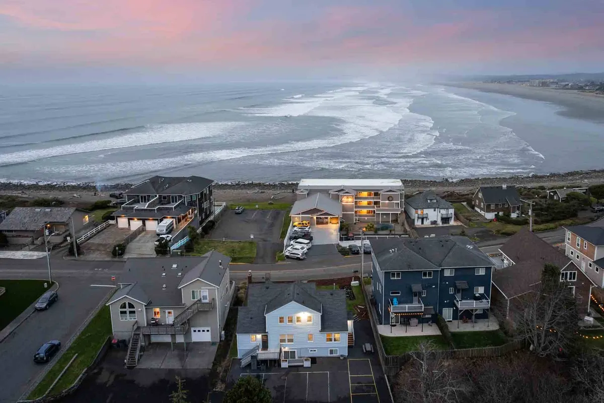 The hero image showcases golden ocean waves rolling onto a sandy shore, with classic beachfront houses lining the coast as the sky glows in vibrant sunset hues. The scene invites visions of leisurely beach walks, family gatherings by the water, and tranquil evenings with the soothing sound of waves in the background.