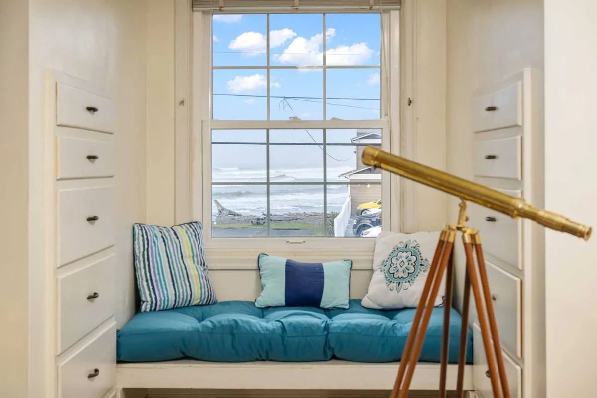 A tranquil interior nook with a cushioned window seat, decorative pillows, a telescope, and a picture window offering a mesmerizing ocean panorama. The space is ideal for quiet moments, reading, or watching the waves roll in.