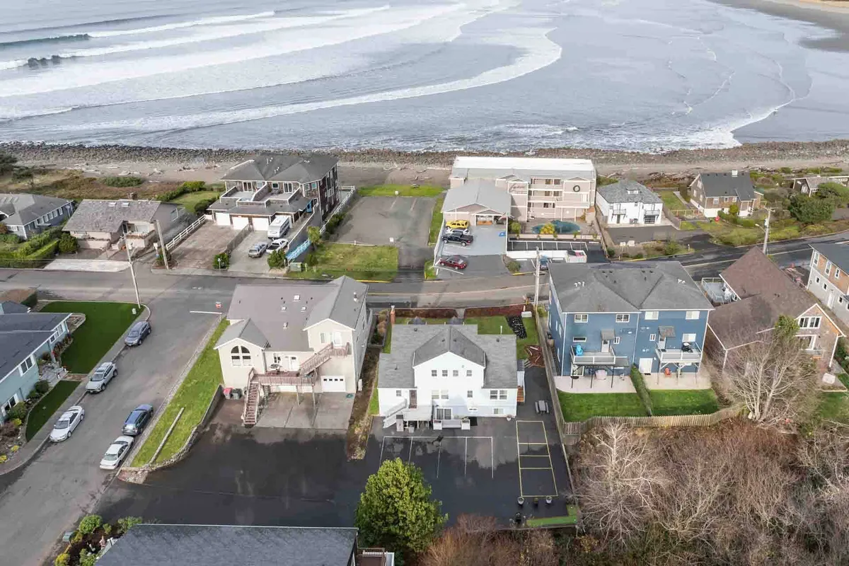 A wide exterior view shows ocean waves rolling onto the sandy beach, with several houses facing the coastline. Green lawns and a road create a welcoming, open feel, while people are seen walking along the beach, surfing, and picnicking, capturing the lively, social atmosphere of coastal exploration.